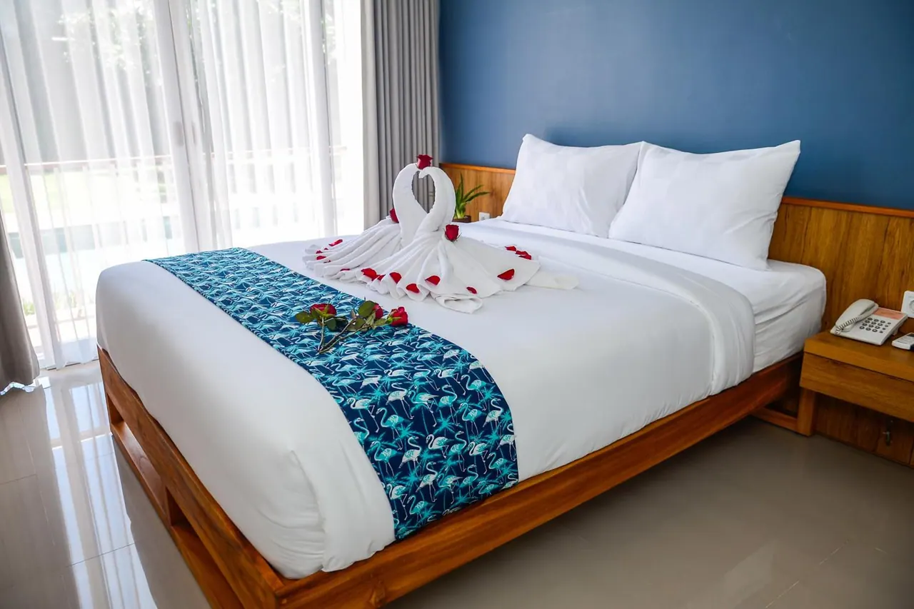 A made-up double bed with white linen, towel swans decorated with roses, and a blue patterned runner sits in front of a blue wall, with a telephone on the bedside table and sheer curtains at the sliding doors.