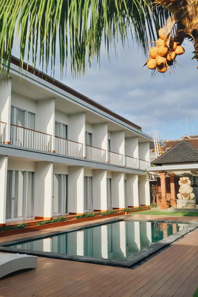 A long rectangular swimming pool on a wooden deck sits in front of a two-storey white building with balconies, with a palm tree and a stone statue at the side.