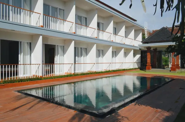A rectangular swimming pool on a timber deck beside a two-storey white building with balconies and a pavilion with red columns on the right.