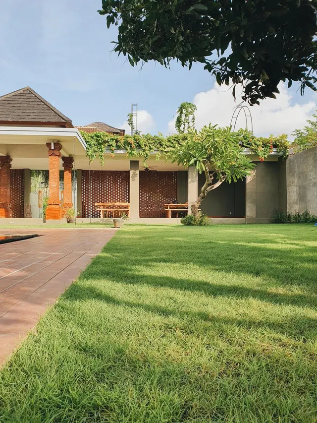 A green lawn with a frangipani tree, a brick-paved path and a covered pavilion with orange columns and wooden benches beside a grey wall.