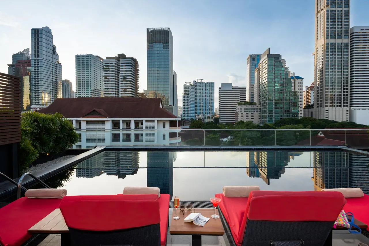 A rooftop infinity-edge pool with red sun loungers and a small table holding drinks and a newspaper, with high-rise towers visible beyond the glass railing.