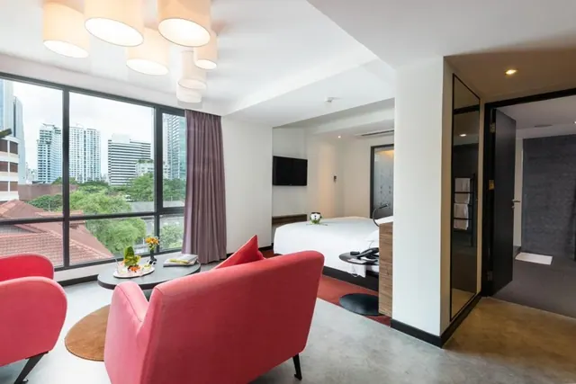 A living area with red armchairs and a round table sits by floor-to-ceiling windows overlooking high-rise buildings, with a bed visible in the adjoining room.