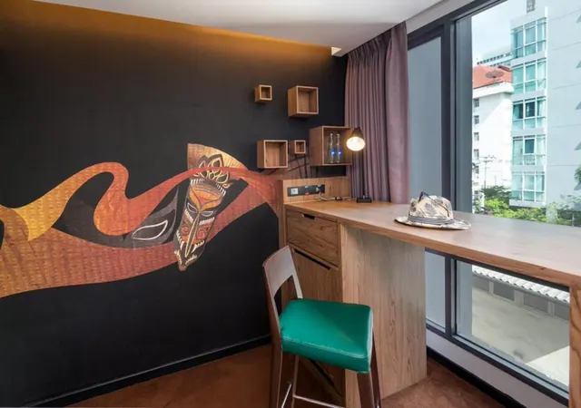 A hotel room with a dark wall painted with a tribal mask mural, a wooden counter desk with a green leather stool, and a window looking onto nearby buildings and trees.