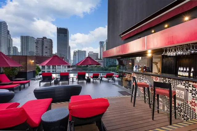 A rooftop terrace with red sun loungers, red parasols and a bar counter with stools, overlooking city high-rises under a partly cloudy sky.