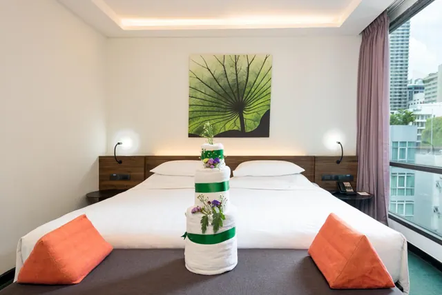 A double bed with two orange cushions and a rolled-towel decoration tied with green ribbon sits in a room with a large window showing city buildings outside.