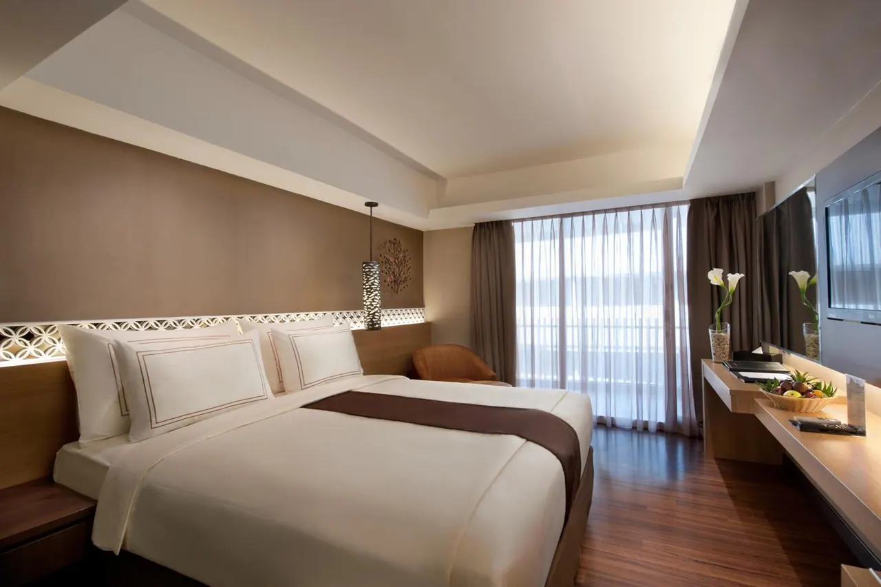 A bedroom with a large bed, an armchair by sheer-curtained windows, a wall-mounted television and a desk with a fruit bowl.