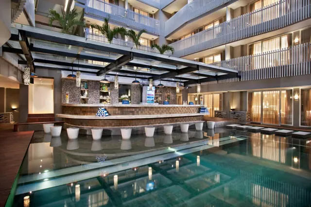 An indoor pool sits beside a bar with stools in the pool, under a multi-storey atrium lined with balconies and sun loungers along the edge.