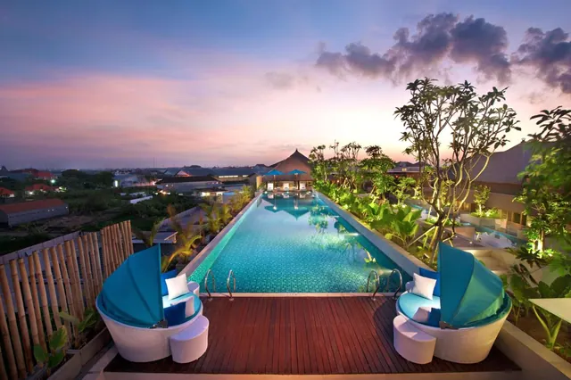 A long illuminated lap pool at dusk on an elevated wooden deck, with blue canopied daybeds and tropical plants along the edge.
