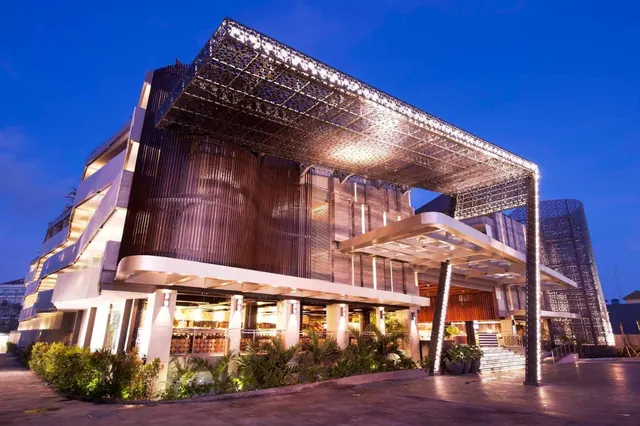 The exterior of a multi-storey building with a perforated lattice canopy and illuminated facade at dusk, with a paved forecourt and planting in front.