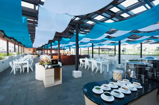An outdoor restaurant with blue canopy shade sails, set tables with white chairs, and a buffet counter with bowls and chafing dishes.