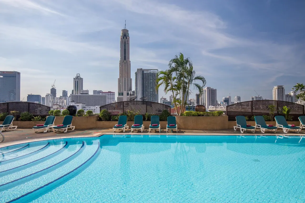 A rooftop swimming pool with turquoise water and teal loungers, with a tall tower and city skyline behind it.