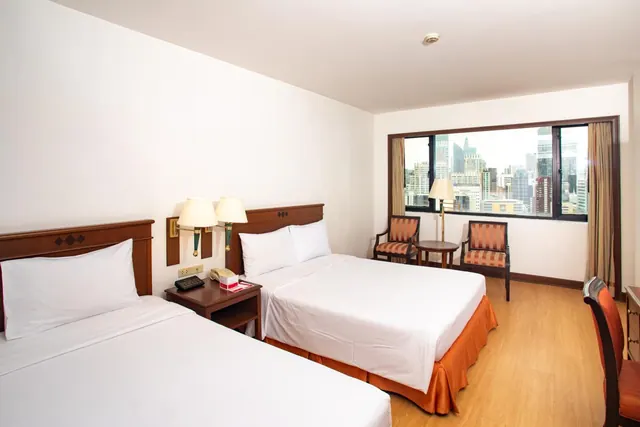 A hotel room with two beds with wooden headboards, a bedside table with a telephone, and two armchairs beside a window looking out over city high-rise buildings.