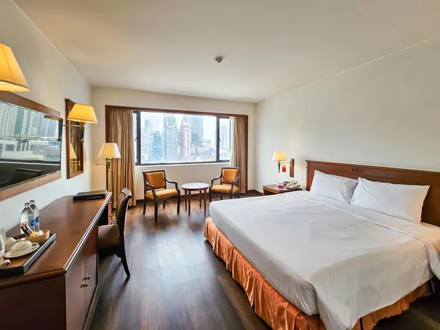 A king bed faces a seating area and a window showing high-rise towers in a hotel bedroom.