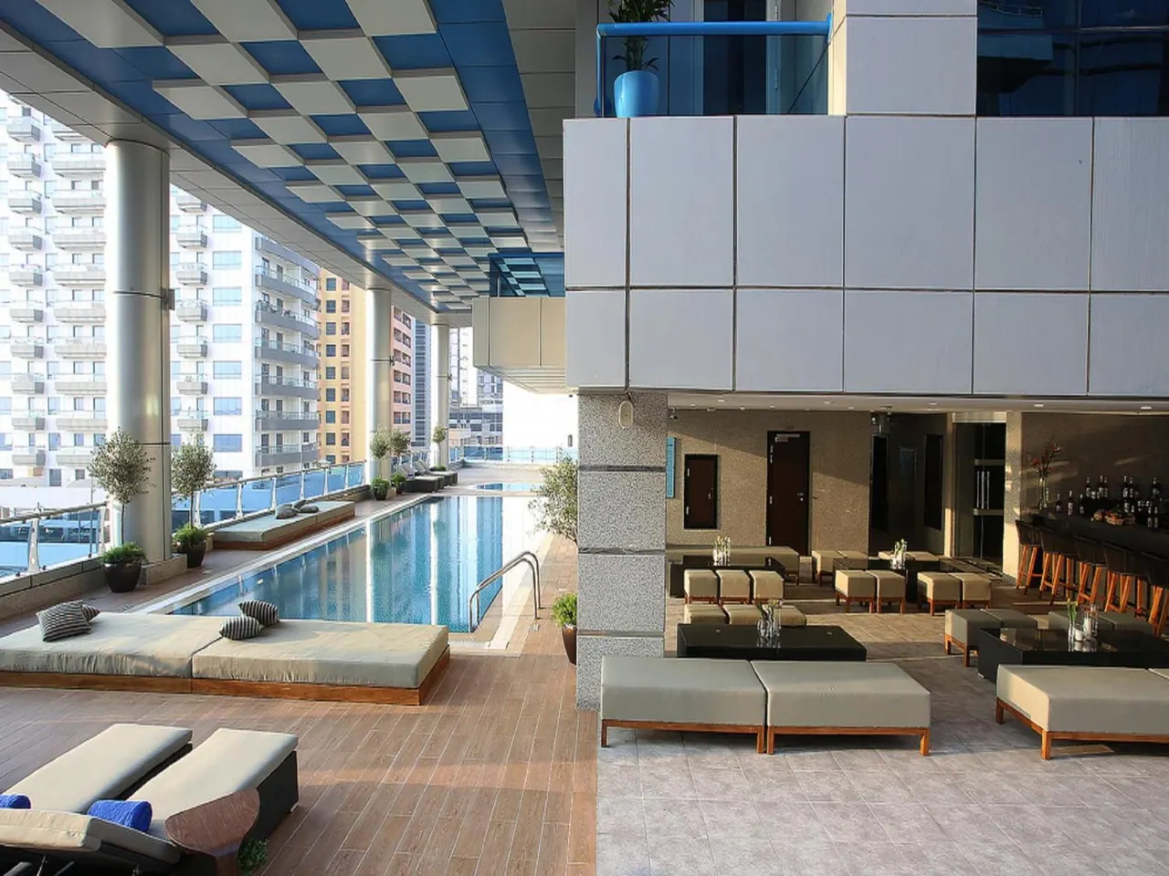 A long rectangular pool sits beside cushioned sun loungers on a covered deck, with low lounge seating and a checkered blue ceiling to the right and residential towers behind.