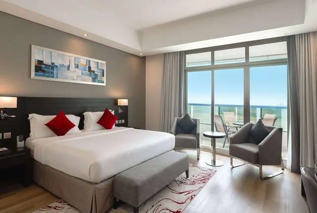 A bedroom with a king bed, two armchairs and a round side table, with floor-to-ceiling glass doors opening onto a balcony with a view of the sea.