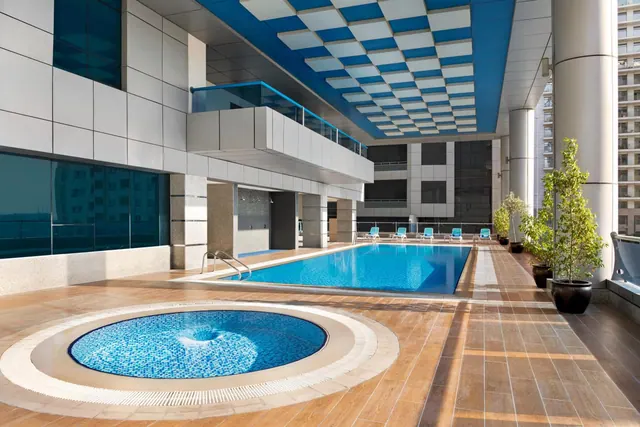 An outdoor pool with a blue checkered ceiling overhead, a round mosaic hot tub in the foreground, blue sun loungers and potted plants along the poolside.