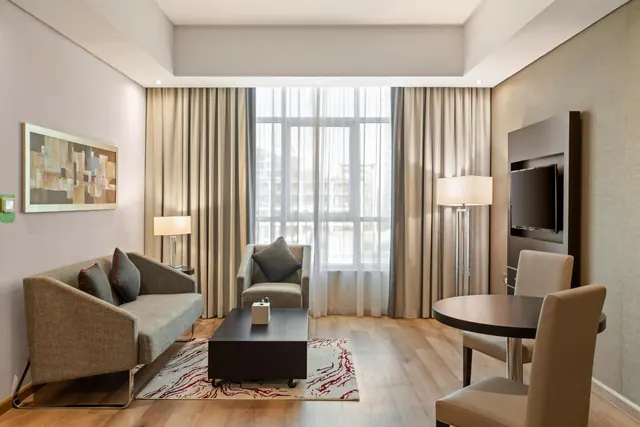 A hotel living room with a grey sofa and armchair, a coffee table, a round dining table with chairs, a wall-mounted television and a large window with sheer curtains.