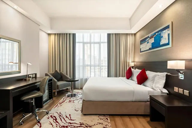 A hotel bedroom with a made double bed with red cushions, a desk and chair, an armchair, a small fridge and tall windows looking onto city buildings.