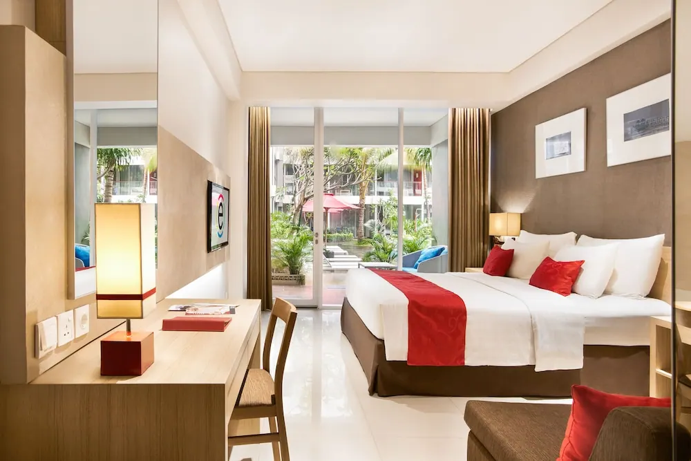 A hotel bedroom with a king bed with red accents, a wooden desk with a lamp, a wall-mounted television, and glass doors opening onto a tropical garden.