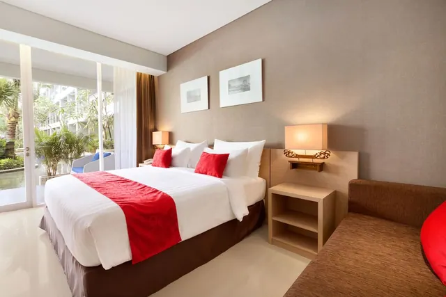 A made king bed with red accents sits beside a brown sofa, with sliding glass doors opening onto a balcony with palm trees outside.