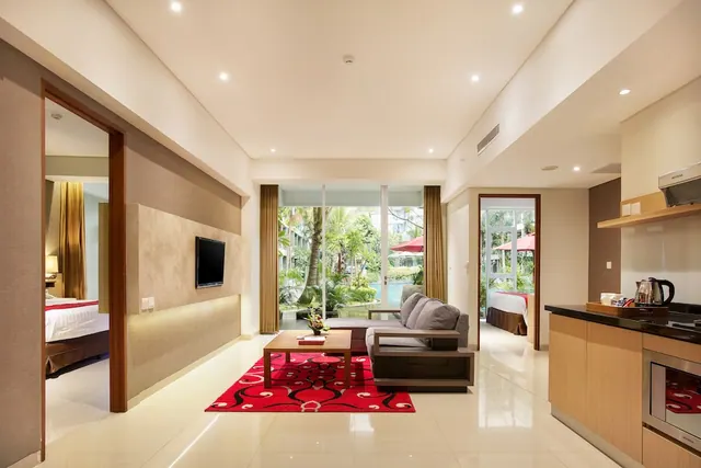 A living area with a grey sofa, wall-mounted television, red rug and wooden coffee table, with a kitchenette on the right and a garden with a pool visible through sliding glass doors.