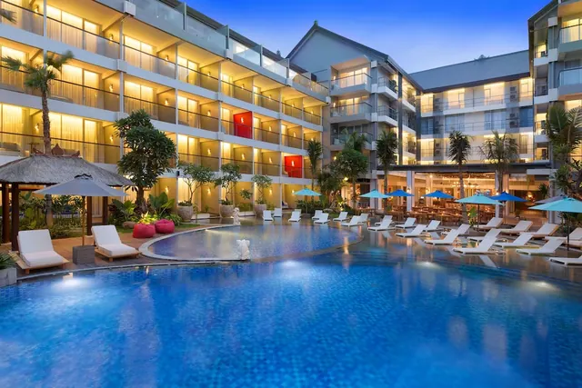 A large illuminated swimming pool at dusk with white sun loungers, blue parasols and hotel balconies behind.