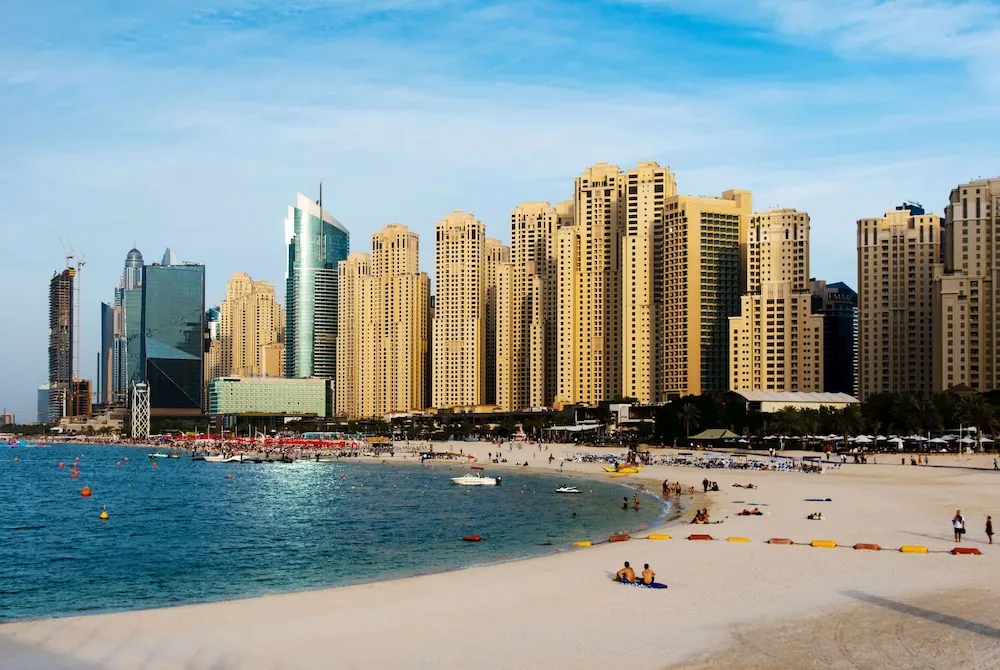 A sandy beach with a few people sitting and walking near the shoreline, with a row of high-rise residential towers behind it under a blue sky.