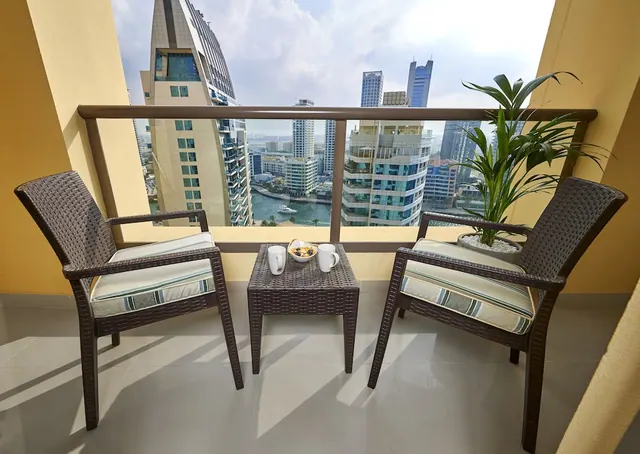 A private balcony with two wicker armchairs, a small table holding two mugs and a bowl, a potted palm, and a view of high-rise towers above a waterway.