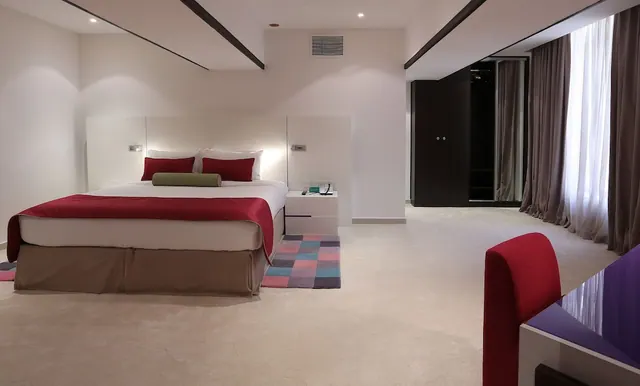 A bedroom with a king bed dressed in red and white bedding, a checkered rug, a dark wardrobe, and a red chair beside a purple desk near a curtained window.