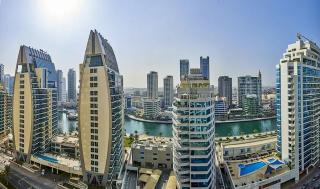 Elevated panoramic view of high-rise towers, a canal with waterside buildings, and a rooftop swimming pool under a hazy sky.