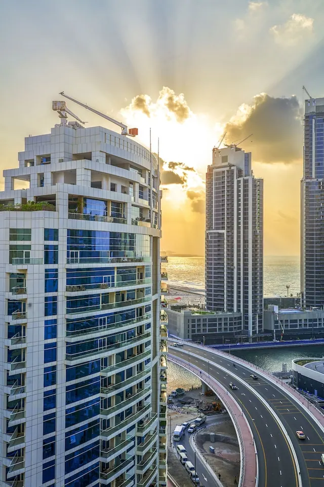 Sunset light over high-rise towers, a curved multi-lane road and the sea, with construction cranes visible on the buildings.