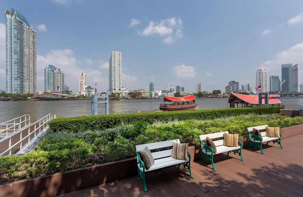 A wooden riverside deck with green-and-white benches and planted borders overlooks a wide river with a red-roofed boat and high-rise towers beyond.