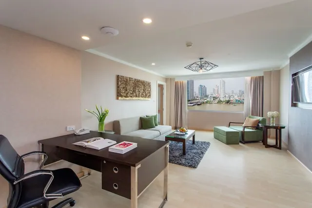 A hotel suite living area with a dark wooden desk, a grey sofa, a green armchair and a large window showing a river and city high-rises.