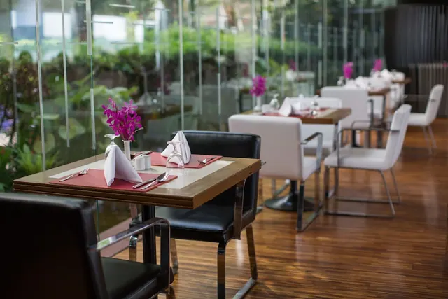 A restaurant dining room with tables set with napkins, cutlery and pink orchids, with glass walls looking onto greenery outside.