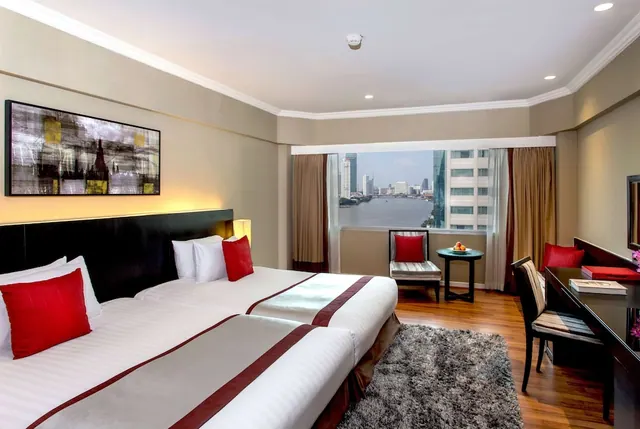 A hotel bedroom with two beds with red cushions, an armchair and a desk, and a window overlooking a river and city buildings.