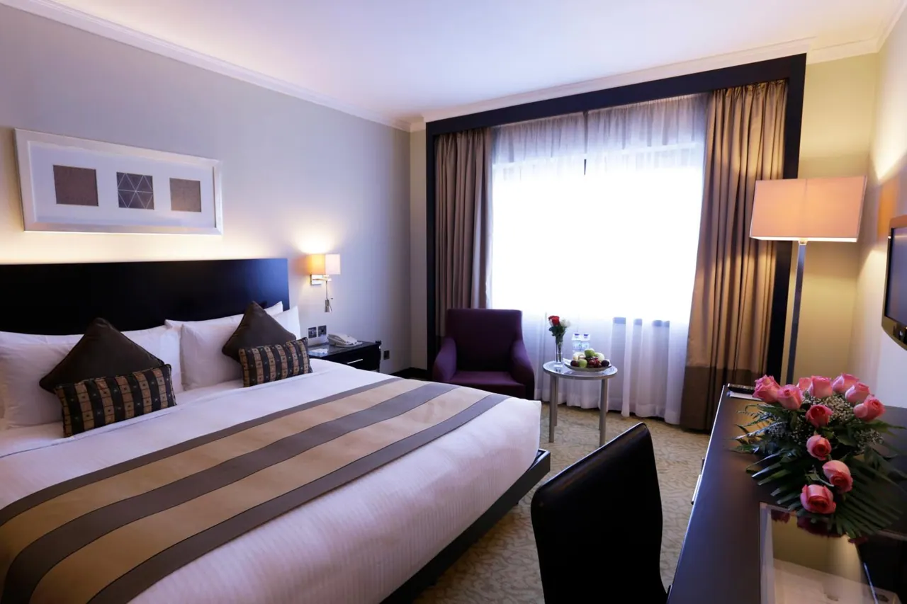 A king bed with a striped runner and cushions sits beside a purple armchair, a round side table with fruit, a desk with roses, and a window with sheer curtains.