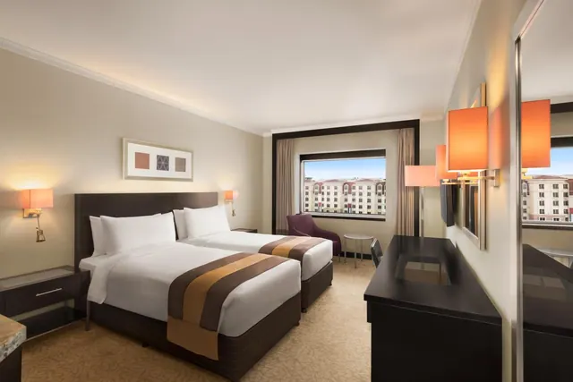 A twin bedroom with two beds, a purple armchair, a wall-mounted television and a window looking out onto multi-storey buildings.
