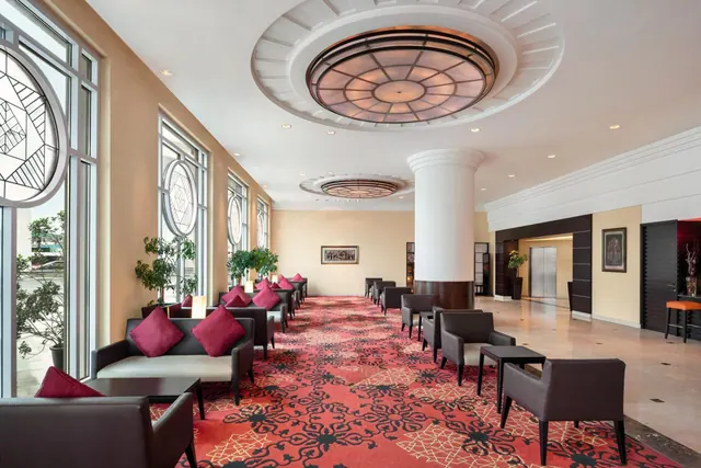 A lounge seating area with dark armchairs, magenta cushions and a red patterned carpet, beside tall windows and a large circular ceiling light.