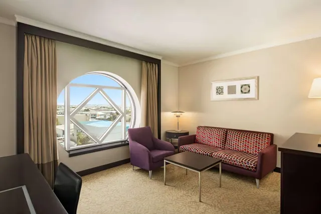 A living area with a round window overlooking rooftops and a pool, a red patterned sofa, a purple armchair, and a low coffee table.