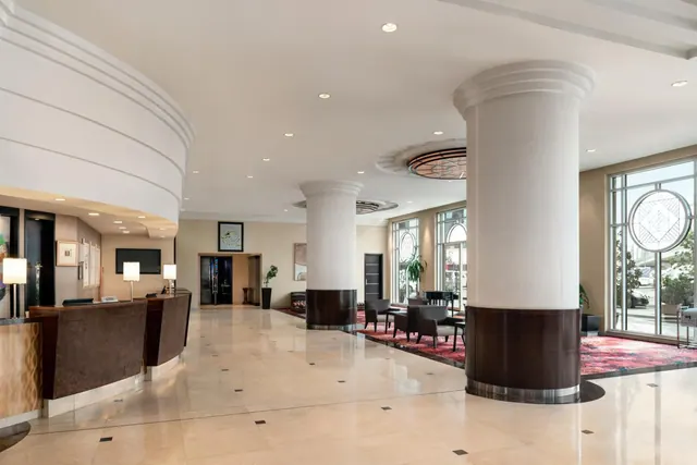 The lobby has a curved reception desk on the left, round white columns, a seating area with armchairs on a red rug, and tall windows on the right.