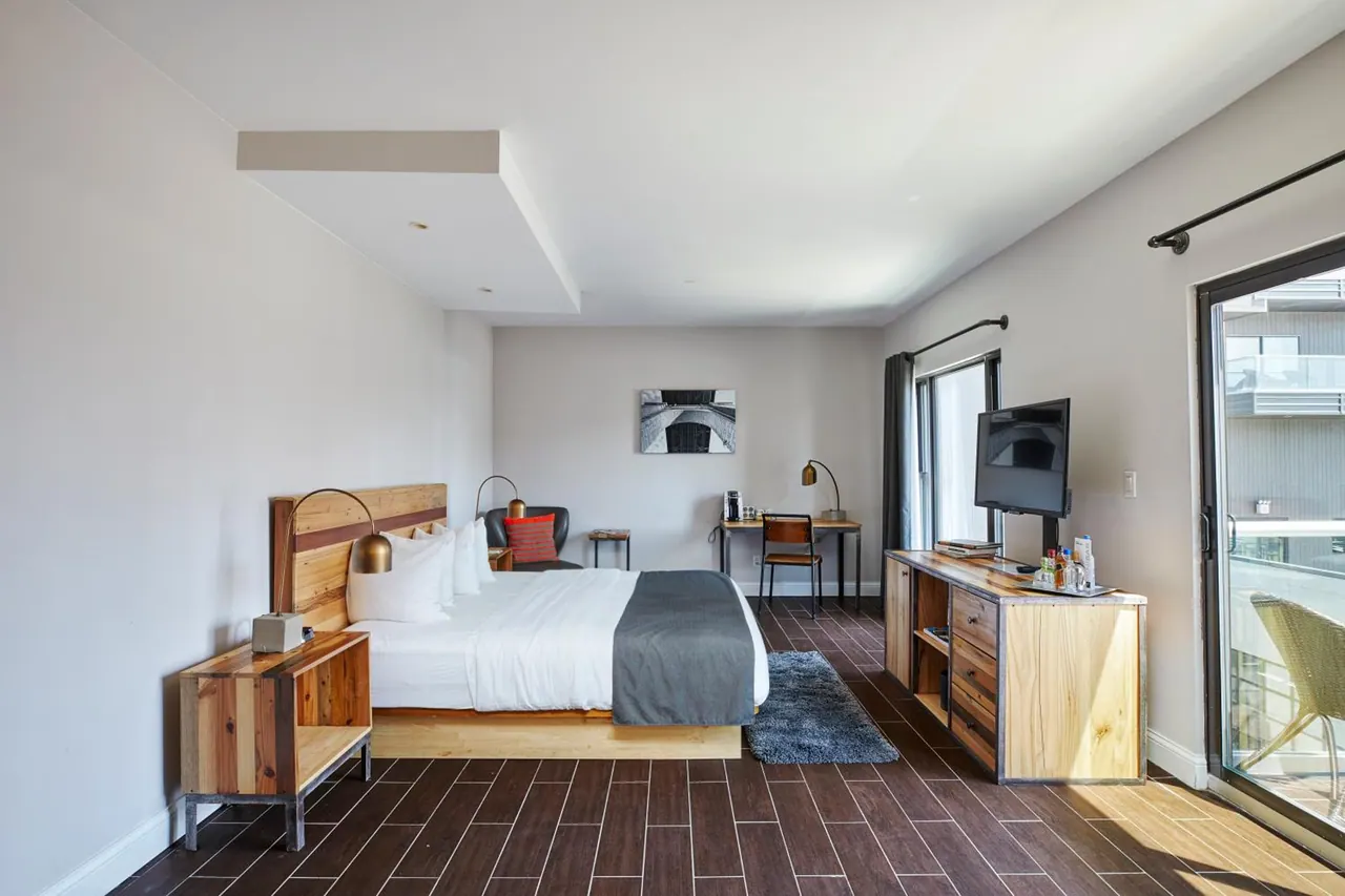 A hotel bedroom with a bed, a wooden dresser with a wall-mounted television, a desk and a sliding door to a private balcony.