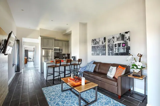 A living room with a brown leather sofa, a wood and metal coffee table on a blue rug, black and white subway train prints on the wall, and an open kitchen with bar stools beyond.