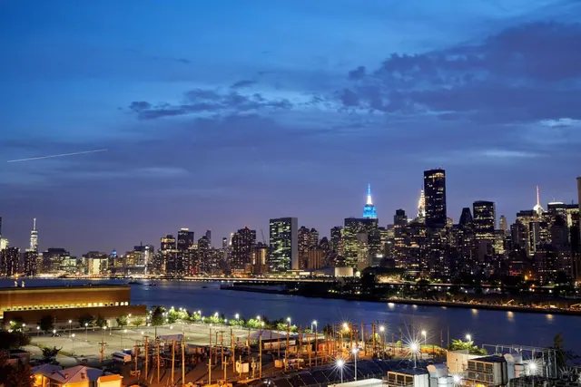Dusk view across the East River toward the illuminated Manhattan skyline, with the Empire State Building lit in blue at centre.