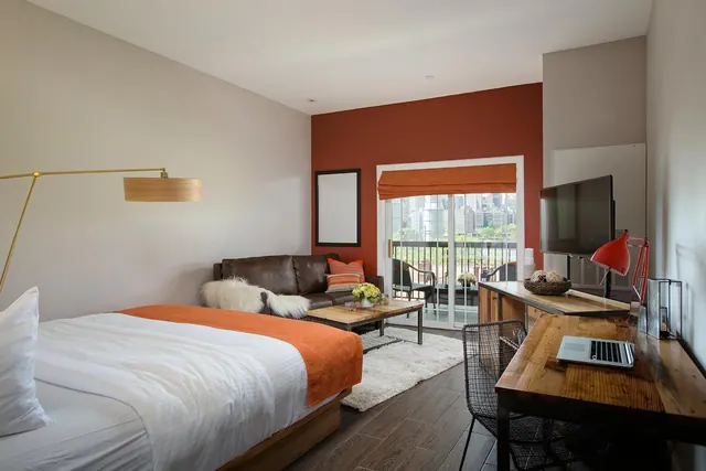 A bedroom with a king bed, brown leather sofa, wooden desk and wall-mounted television, with a glass sliding door opening onto a balcony with a city skyline view.
