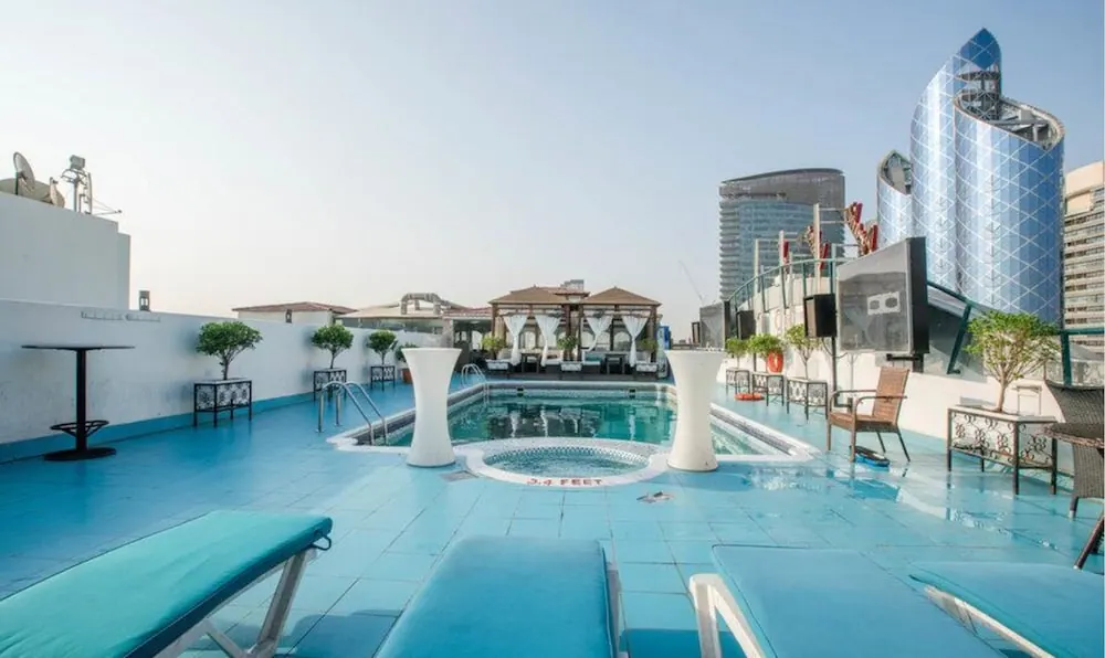 A rooftop pool with a round hot tub, blue tiled deck, sun loungers and tall glass towers in the background.