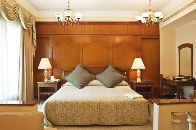 A bedroom with a double bed under a patterned cover, a wood-panelled headboard wall, bedside lamps, two chandeliers and a desk with a mirror.