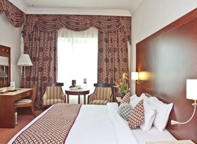A hotel bedroom with a made double bed, a wooden headboard, a desk, two striped armchairs and a round side table in front of a curtained window.