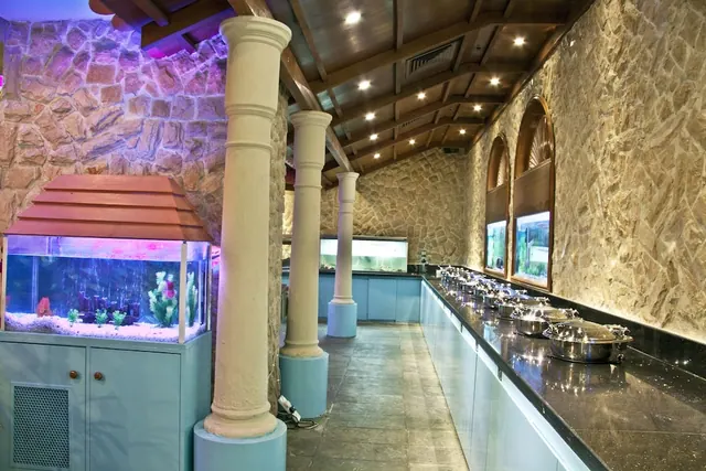 A restaurant buffet counter with covered chafing dishes runs along a stone wall, beside a built-in aquarium and stone-clad columns.