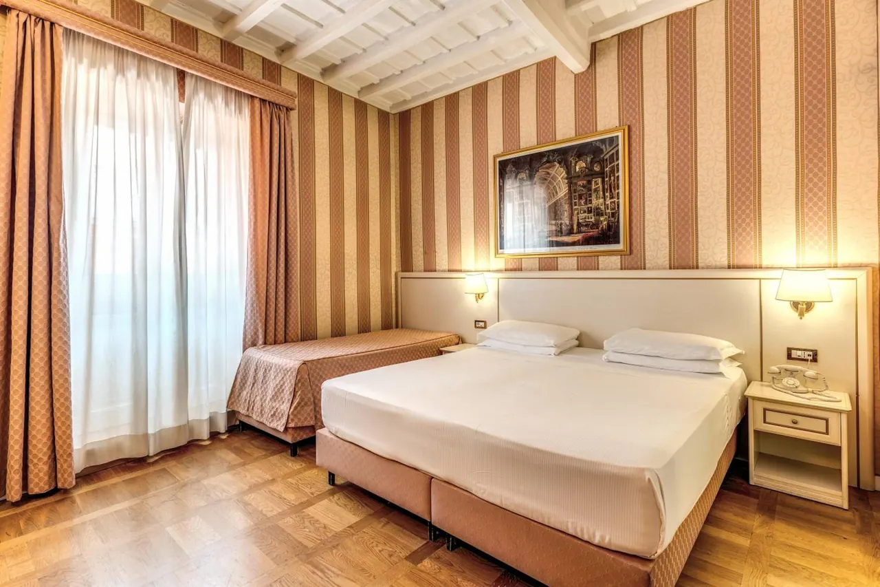 A bedroom with a double bed, a single extra bed beside the sheer-curtained window, striped wallpaper, a framed painting and a wooden parquet floor.