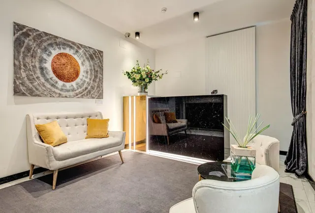A lounge seating area with a white tufted sofa, yellow cushions, round white armchairs, a glass table and a large abstract painting on the wall.